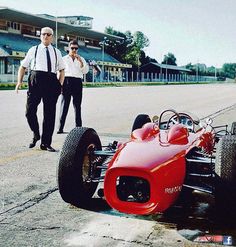 a man standing next to a red race car on top of a tarmac with another man in the background