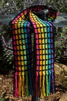 a multicolored crocheted shawl hanging on a pole in front of some bushes