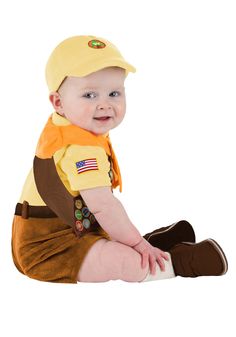 a baby sitting on the ground wearing a yellow hat and brown pants with an american flag patch