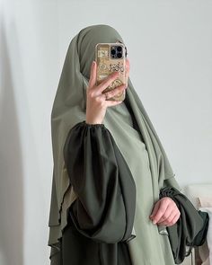 a woman wearing a hijab taking a selfie with her cell phone while standing in front of a white wall