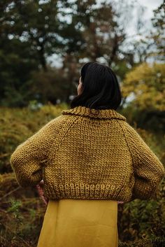 a woman standing in the woods with her back to the camera, wearing a mustard colored coat