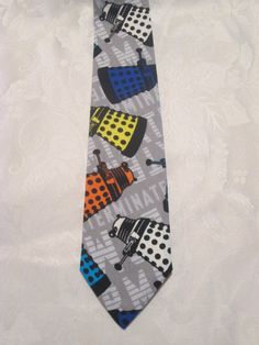 "What Whovian could resist a colorful Dalek necktie? White, orange, yellow, indigo, and turquoise Daleks cascade across a pale grey background (which features the word \"Exterminate!\" in white). Appearance of Daleks will vary slightly on the tie due to pattern placement.  This is the perfect for when you \"have to wear a tie\". Handmade of 100% cotton in Massachusetts. Tie is 53\" long and approximately 2 inches wide at the widest point." Dr Who Dalek, Doctor Who Dalek, Bbc Doctor Who, Timey Wimey Stuff, Grey Background, Daughter Of God, Tie Accessories, Dr Who, Suit And Tie