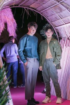 two men standing next to each other in front of a bamboo hut with pink carpet