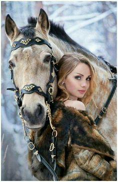 a beautiful young woman standing next to a brown and black horse in the snow with it's bridle on