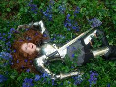 a woman dressed in armor laying on the ground surrounded by flowers and bluebells