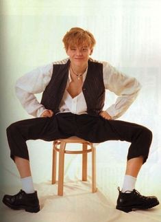 a man sitting on top of a wooden chair next to a white wall with his legs crossed