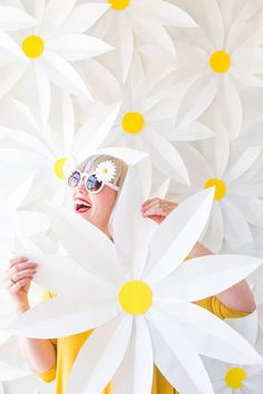 a woman in yellow dress and sun glasses holding up large white paper flowers with yellow dots on them