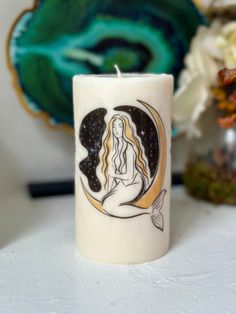 a white candle with a drawing of a mermaid sitting on it's side next to flowers