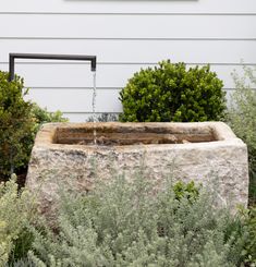 a water fountain in front of a house with bushes and shrubbery around the corner