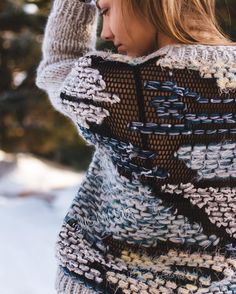 a woman wearing a knitted sweater and holding her hand up to her head while standing in the snow