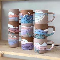 four coffee mugs sitting on top of a wooden shelf with mountains and clouds painted on them