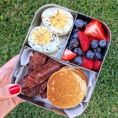 a person holding up a lunch box filled with bacon, eggs, pancakes and fruit