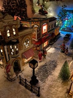 a toy town is shown in the snow at christmas time with lights on and people walking around