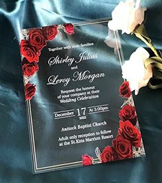 a wedding card with roses on it sitting on a blue satin covered bed sheet in front of a bouquet of white and red flowers