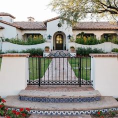 an iron gate in front of a white stucco house with red flowers and greenery