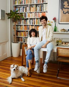 a man and woman sitting on a chair in front of a bookshelf with a dog