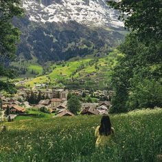 a woman sitting in the grass looking down at a village