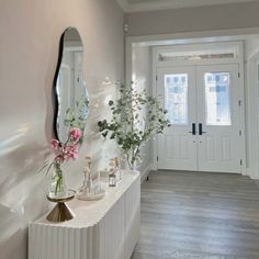 a white hallway with vases and flowers on the side table next to a mirror