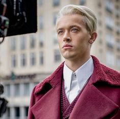 a man with blonde hair wearing a red coat in front of a camera and buildings