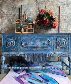 an old blue painted dresser with fringes and flowers on the top, in front of a brick wall