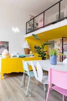 a dining room with yellow and pink chairs, white table and bright colored cabinet in the background