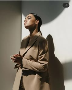 a woman standing in front of a white wall wearing a tan coat and black pants