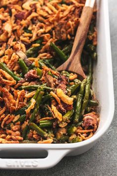 a casserole dish filled with green beans, bacon and other toppings next to a wooden spoon