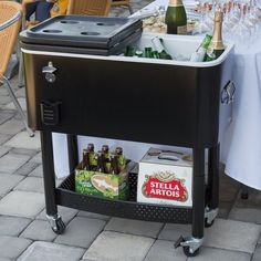 an outdoor bar cart with beer bottles and cans on it's wheels, next to a table full of wine glasses