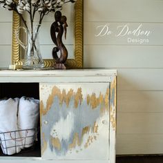 a white cabinet with gold paint on it and some flowers in a vase next to it