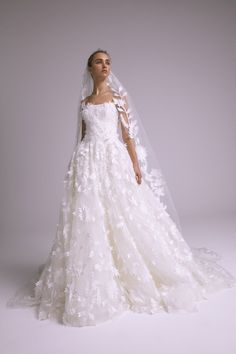 a woman in a white wedding dress with flowers on the skirt and veil, standing against a gray background