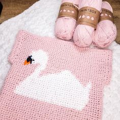 two balls of yarn are next to a pink crocheted swan blanket and ball of yarn