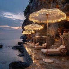 several lounge chairs and umbrellas are lit up by lights on the rocks next to the ocean