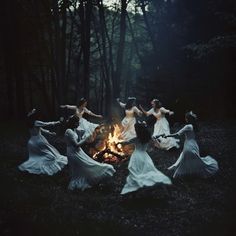 four women in white dresses are dancing around a campfire