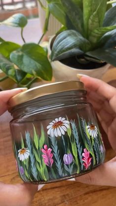 two hands holding a jar with flowers painted on it next to a potted plant