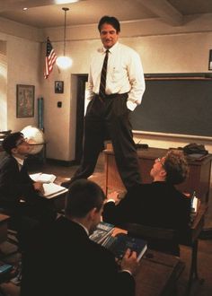 a man standing on top of a desk in front of other men sitting at desks