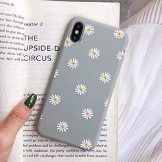 a woman's hand holding up a phone case with daisies on it, next to an open book