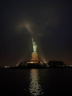 the statue of liberty is lit up at night with its lights on and reflecting in the water