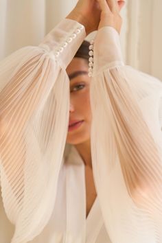 a woman with her hands on her head, wearing a white blouse and pearl earrings