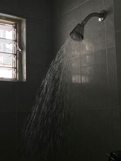 a shower head with water coming out of it in a black tiled bathroom next to a window