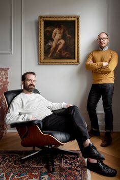 two men standing next to each other in front of a painting and an eames chair