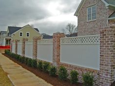 a brick fence is lined with bushes and shrubs in front of a house on a cloudy day