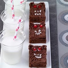 three brownies with chocolate frosting and candy eyes are on a plate next to two glasses of milk