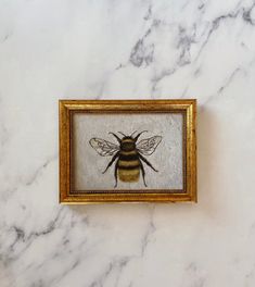 a framed painting of a bee on a white marble counter top with gold trimming