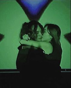 two women hugging each other in front of a green wall with blue lights on it