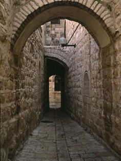 an alley way with stone walls and arched doorways, leading to the light at the end