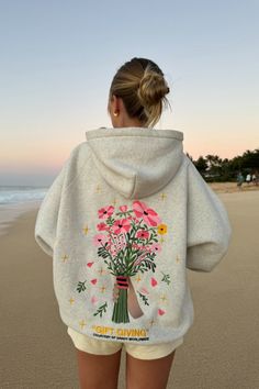 photo shows a blonde teenage girl standing on a sandy beach at sunset. she is facing away from the camera, looking at the ocean in the distance. she is wearing cream colored sweat shorts and a gray hoodie that has a big embroidered graphic of a bouquet of flowers on the back. "gift giving" and "courtesy of dandy worldwide" are embroidered on the back as well. the girl has her hair in a bun. the sky is orange and blue, like the sun is just about to set Dandy Flower Hoodie, Dandy Hoodie Gift Giving, Gift Giving Hoodie, Danny Worldwide Hoodie, Cute Aesthetic Hoodies, Cute Things To Ask For Christmas, Hoodies That Hoodie, Embroidery Designs On Hoodies, Dandy Worldwide Hoodie