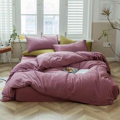 a bed with pink sheets and pillows in front of two windows that have white shutters