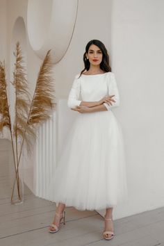 a woman standing in front of a wall wearing a white dress and heels with her arms crossed