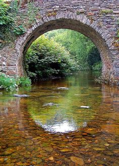 an arched stone bridge over a river with clear water and rocks on the ground below