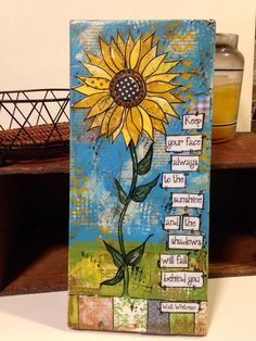 a painting with words written on it and a sunflower in the middle, sitting on a shelf